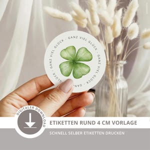 May include: A circular sticker featuring a watercolor illustration of a green four-leaf clover in the center. The text GANZ VIEL GLÜCK is printed in a circular pattern around the clover. The sticker has a white background and a smooth edge.