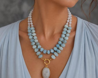 Puede incluir: Un collar de varias hebras con cuentas facetadas en rosa pálido y azul claro. El collar incluye un cierre circular dorado que conecta con un gran colgante de piedra blanca de forma ovalada y lisa. El diseño incorpora pequeños espaciadores dorados.