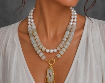 Puede incluir: Un collar de dos hileras con perlas esféricas blancas y cuentas cilíndricas de piedra gris claro. El collar incluye un cierre circular dorado en el centro, del cual cuelga un gran colgante de ágata irregular y translúcida en tonos marrón y blanco.