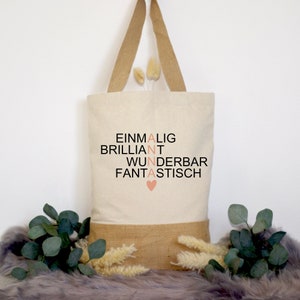 Könnte beinhalten: Eine weiße Canvas-Tragetasche mit braunen Jutegriffen und einem braunen Juteboden. Die Tasche hat schwarzen Text, der "EINMALIG BRILLIANT WUNDERBAR FANTASTISCH" mit einem rosa Herzen in der Mitte lautet.