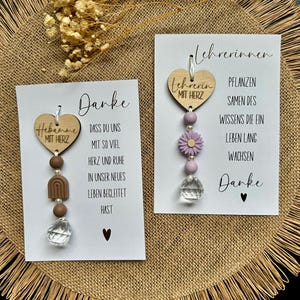 Op de afbeelding: Twee witte kaarten met houten hartvormige ornamenten. Op de ene staat "Hebamme MIT HERZ" en "Danke". Op de andere staat "Lehrerin MIT HERZ" en "Danke". De ornamenten hebben kralen, een bloem en een kristal. De achtergrond is een jute textuur.