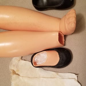 May include: Doll parts including two plastic legs, a pair of black shoes, and a white sock. The legs are a light skin tone, and the shoes are black with a rounded toe. The sock is off-white.