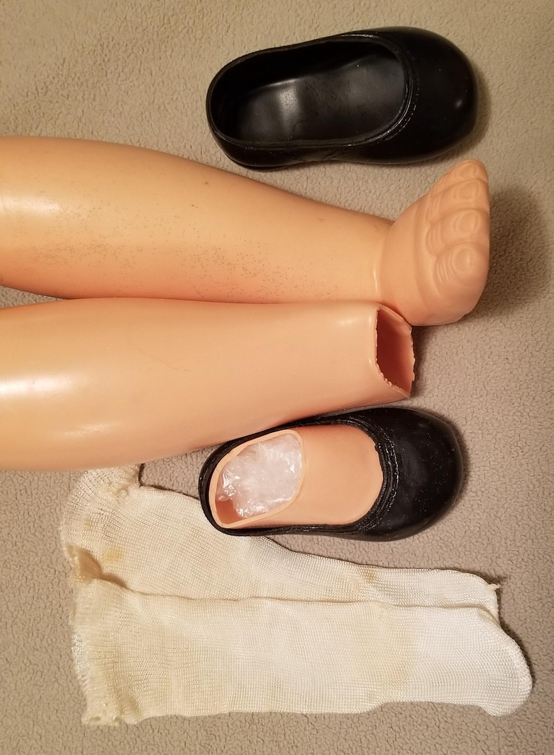 May include: Doll parts including two plastic legs, a pair of black shoes, and a white sock. The legs are a light skin tone, and the shoes are black with a rounded toe. The sock is off-white.
