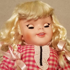 May include: A vintage doll with closed eyes and a gentle smile. The doll has blonde, curly hair and is wearing a pink and white checkered dress with a white lace collar. Pink ribbons adorn the hair and dress.