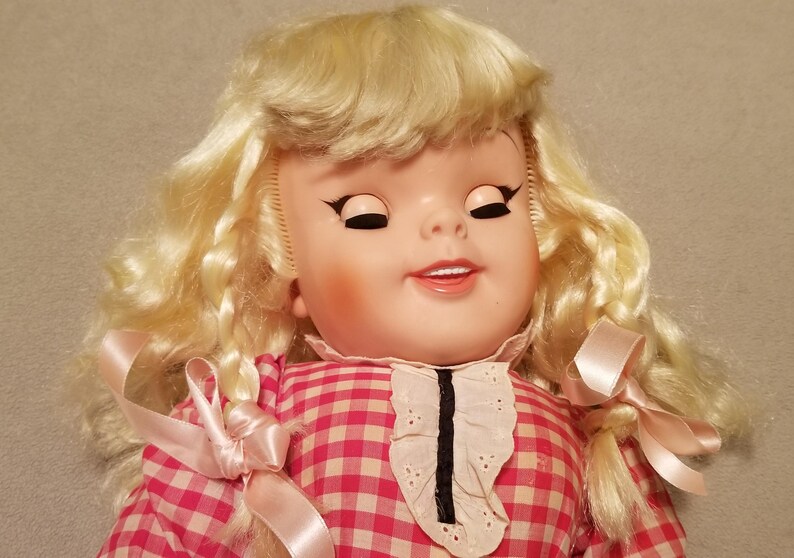 May include: A vintage doll with closed eyes and a gentle smile. The doll has blonde, curly hair and is wearing a pink and white checkered dress with a white lace collar. Pink ribbons adorn the hair and dress.
