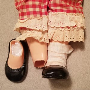 May include: A doll's legs and feet are visible, wearing red and white checkered pants with cream-colored lace trim. The doll has a black shoe and a white sock. The doll's legs are a light peach color.
