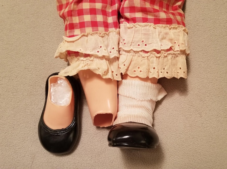 May include: A doll's legs and feet are visible, wearing red and white checkered pants with cream-colored lace trim. The doll has a black shoe and a white sock. The doll's legs are a light peach color.