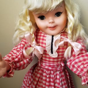 May include: A vintage doll with blonde hair styled in braids and bangs. The doll wears a red and white checkered dress with a white collar and pink bows. The doll has black eyeliner and a slight smile.