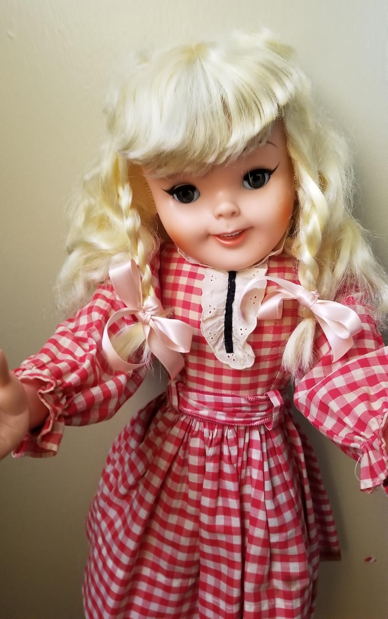 May include: A vintage doll with blonde hair styled in braids and bangs. The doll wears a red and white checkered dress with a white collar and pink bows. The doll has black eyeliner and a slight smile.