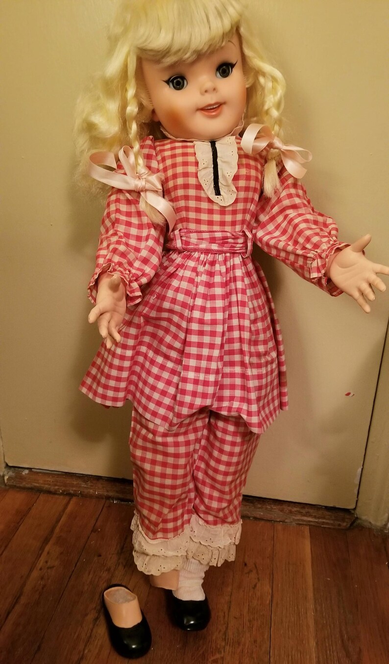 May include: A vintage doll with blonde curly hair, wearing a red and white gingham dress, pants, and a matching belt. The doll has black shoes and a pair of shoes on the floor. The doll's outfit has white lace trim.
