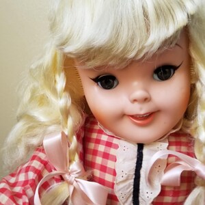 May include: A vintage doll with blonde hair styled in braids and a red and white checkered dress. The doll has large, dark eyes with long eyelashes and a gentle smile. Pink ribbons adorn the braids and dress.