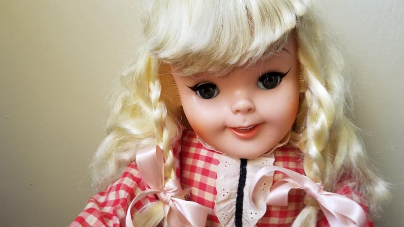 May include: A vintage doll with blonde hair styled in braids and a red and white checkered dress. The doll has large, dark eyes with long eyelashes and a gentle smile. Pink ribbons adorn the braids and dress.