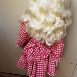 May include: A vintage doll with long, curly blonde hair and a red and white checkered dress. The dress features long sleeves and a bow at the back. The doll's legs are visible, and the overall aesthetic is reminiscent of classic children's toys.