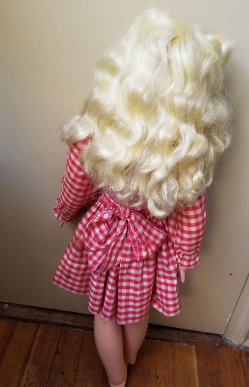 May include: A vintage doll with long, curly blonde hair and a red and white checkered dress. The dress features long sleeves and a bow at the back. The doll's legs are visible, and the overall aesthetic is reminiscent of classic children's toys.