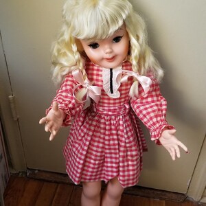 May include: A vintage doll with blonde curly hair and a red and white checkered dress. The doll has a white collar with a black trim and pink bows. The doll is standing on a wooden floor.