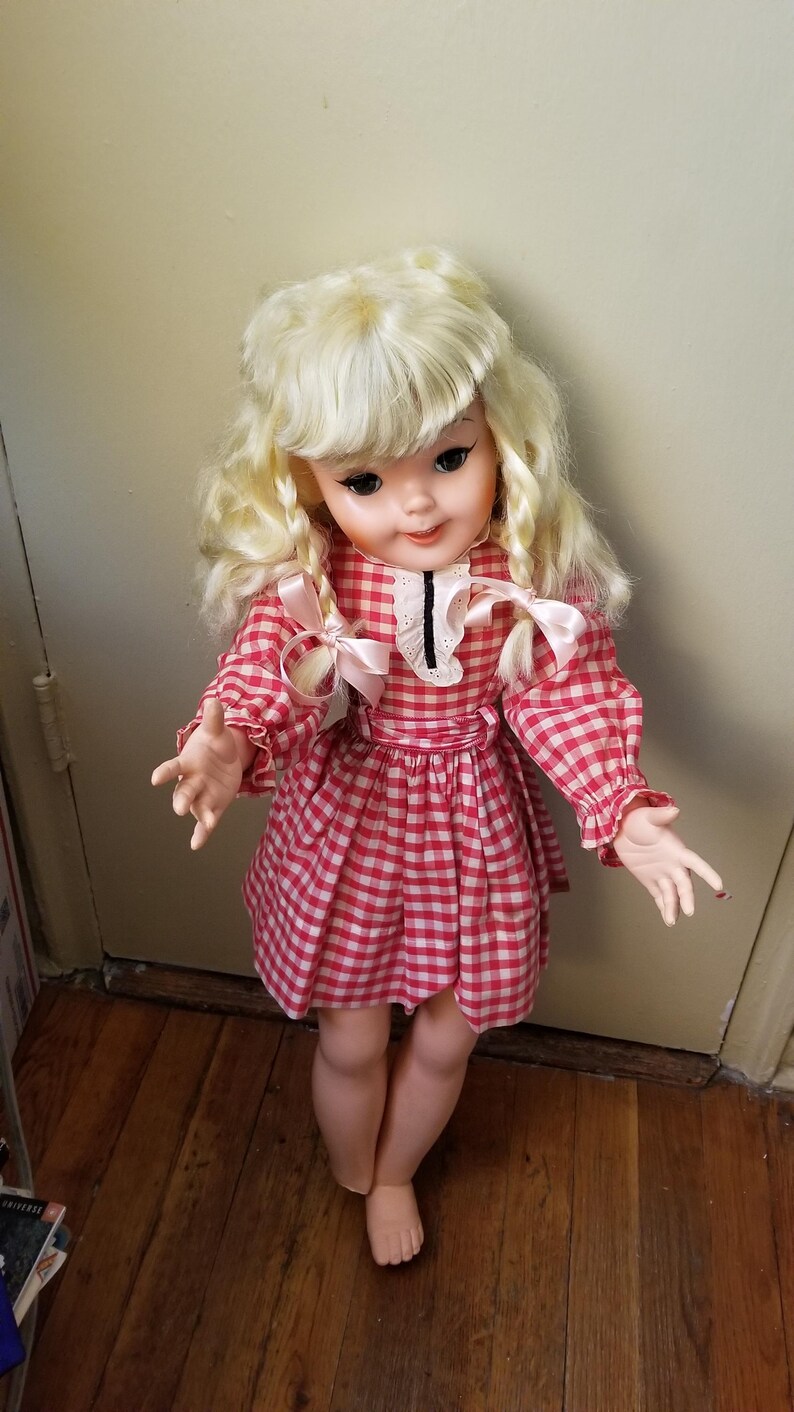 May include: A vintage doll with blonde curly hair and a red and white checkered dress. The doll has a white collar with a black trim and pink bows. The doll is standing on a wooden floor.