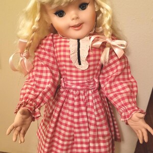 May include: A vintage doll with blonde hair styled in ringlets and bangs. The doll wears a red and white checkered dress with a white collar and pink bows. The doll has blue eyes and is standing on a wooden floor.