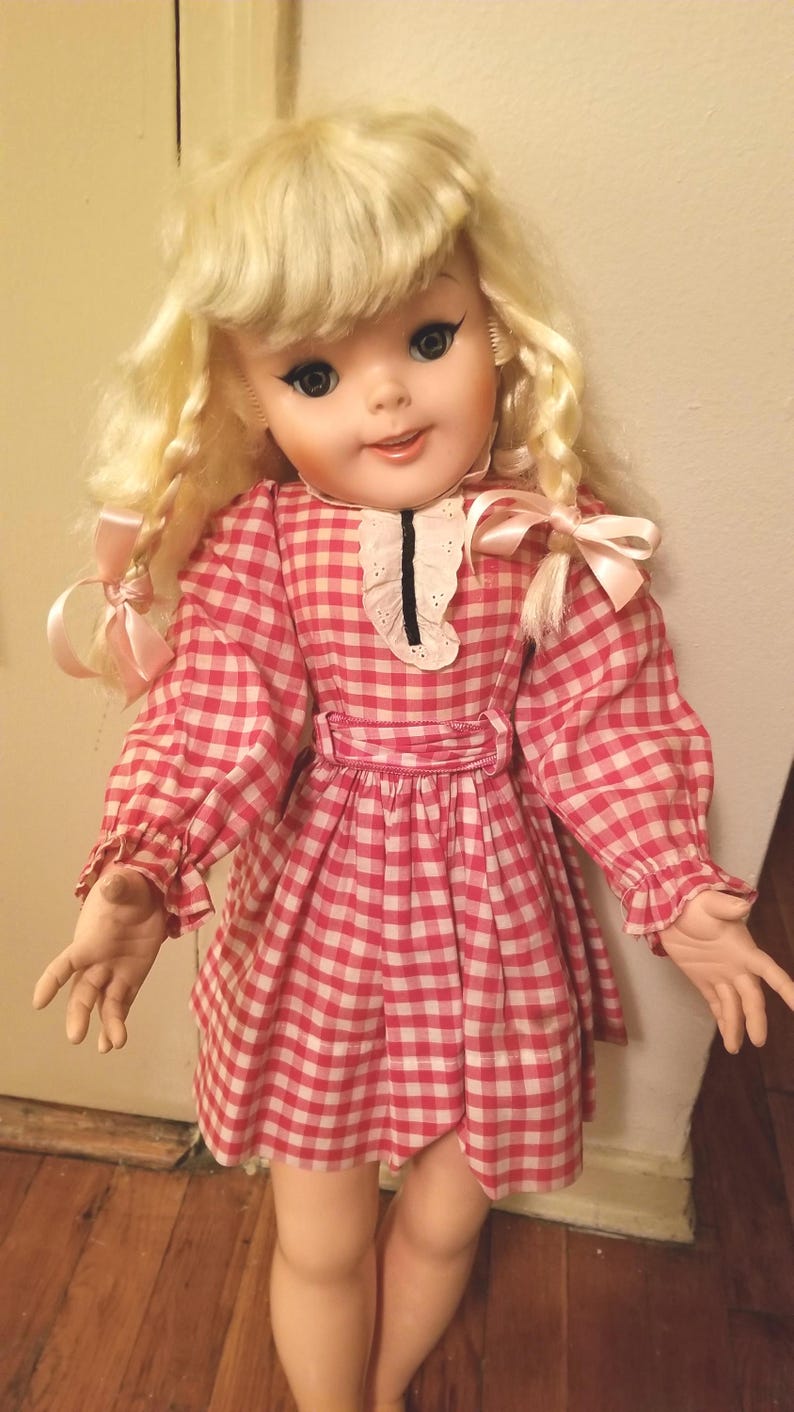 May include: A vintage doll with blonde hair styled in ringlets and bangs. The doll wears a red and white checkered dress with a white collar and pink bows. The doll has blue eyes and is standing on a wooden floor.