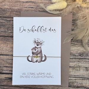 Könnte beinhalten: Eine Grußkarte mit einer Aquarellillustration von zwei sich umarmenden Ottern, die mit Blumenkränzen geschmückt sind. Die Karte trägt die Aufschrift "Du schaffst das" und "Viel Stärke, Wärme und ein Herz voller Hoffnung". Ein goldfarbener Herzanhänger ist befestigt.