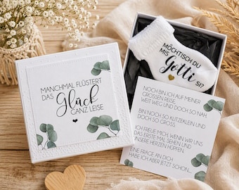May include: A white gift box with a lid featuring the text "MANCHMAL FLÜSTERT DAS GLÜCK GANZ LEISE" and eucalyptus leaves. Inside, a white baby sock displays "MÖCHTISCH DU MIS GOTTI SI?" with a heart. A card includes German text and eucalyptus.