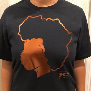 Könnte beinhalten: Ein dunkelblaues T-Shirt mit einer kupferfarbenen Silhouette einer Person mit Afro. Der Text "BS" ist in einem kupferfarbenen Rechteck unten rechts auf dem T-Shirt gedruckt.