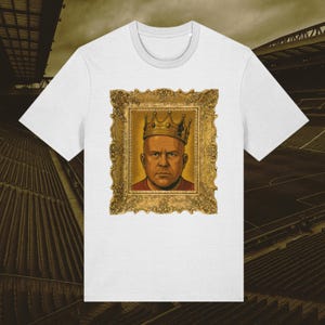 Può includere: T-shirt bianca con un ritratto incorniciato di un uomo con una corona. Il ritratto ha una cornice dorata e uno sfondo giallo. L'uomo nel ritratto ha un'espressione seria. La t-shirt è realizzata in un materiale morbido.