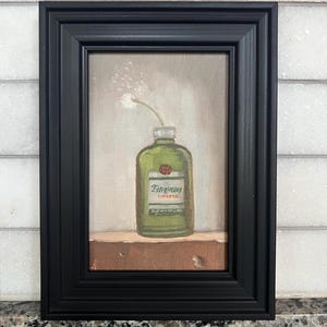 May include: A framed painting featuring a green gin bottle with the Tanqueray label and a single white flower. The artwork is set against a neutral background and framed in a black, rectangular frame.