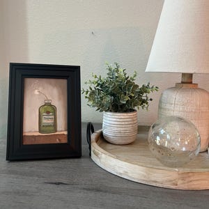 May include: A framed artwork featuring a green bottle with the word "Tincture" on it, a small potted plant, a decorative lamp, and a glass sphere. The items are arranged on a wooden tray.