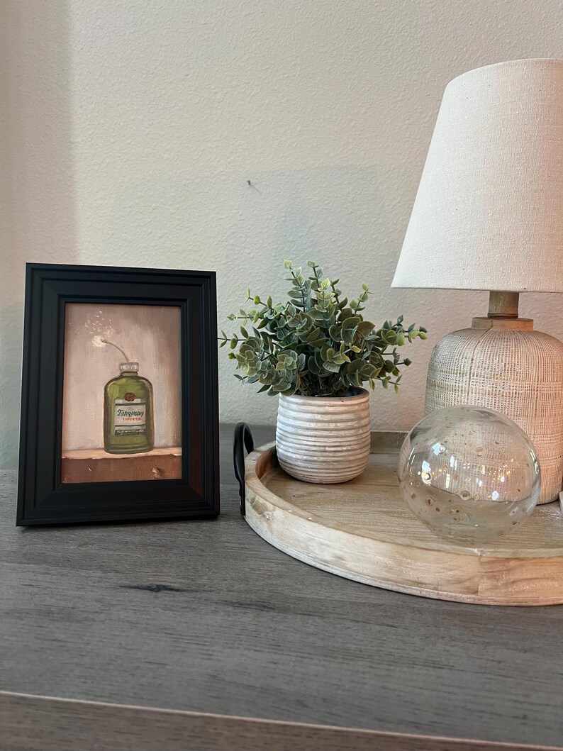 May include: A framed artwork featuring a green bottle with the word "Tincture" on it, a small potted plant, a decorative lamp, and a glass sphere. The items are arranged on a wooden tray.