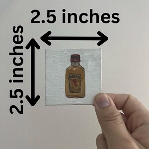 以下が含まれることがあります： 赤いキャップと茶色のラベルが付いた酒瓶の小さな正方形の絵。絵は白いキャンバスに描かれており、サイズは6.35 cm x 6.35 cmです。絵は白い背景に対して手で持たれています。
