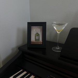 May include: A framed painting of a green bottle with a white flower, next to a martini glass with a lemon twist. The items are on a black piano. The painting is in a black frame.