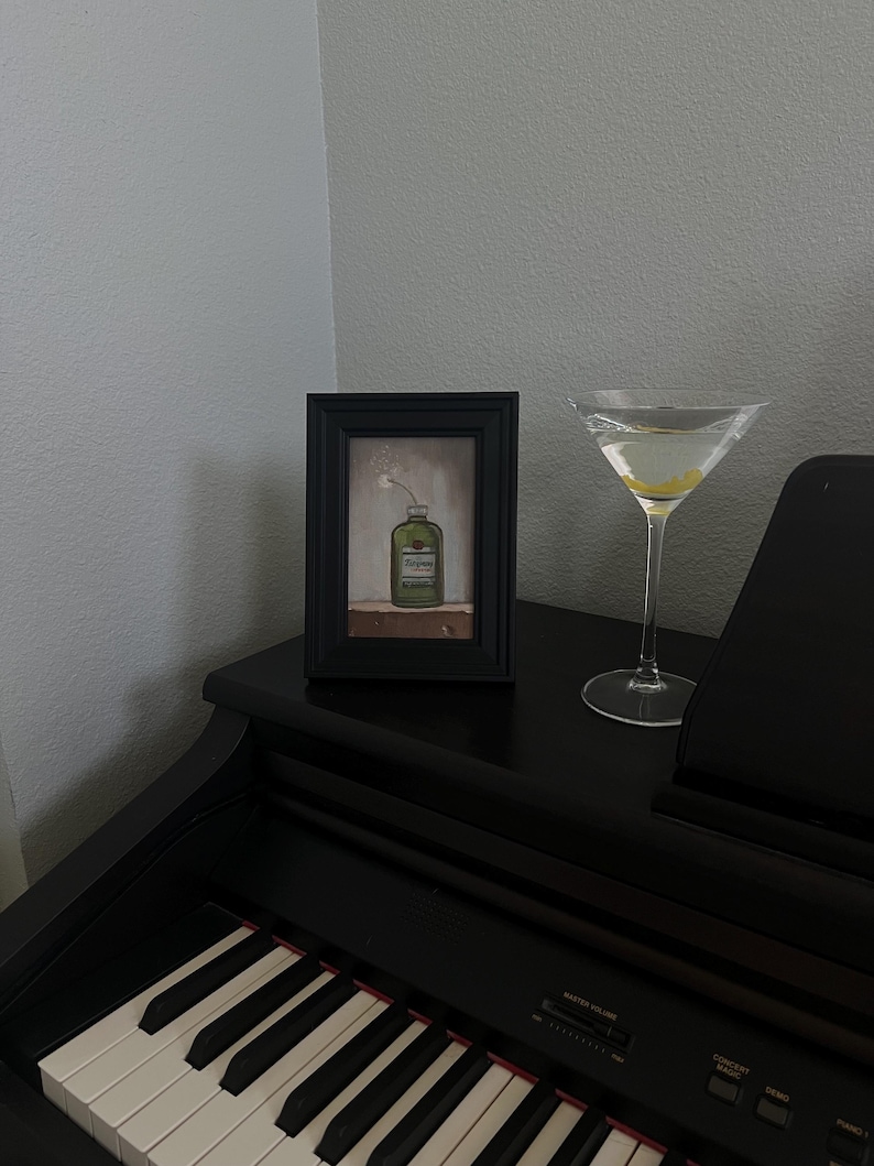 May include: A framed painting of a green bottle with a white flower, next to a martini glass with a lemon twist. The items are on a black piano. The painting is in a black frame.