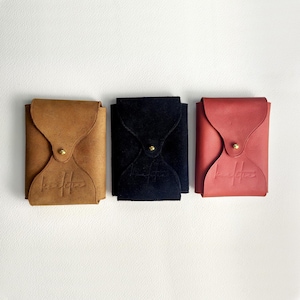Puede incluir: Tres carteras de cuero en color marrón, negro y rojo, dispuestas una al lado de la otra. Cada cartera tiene un cierre de solapa asegurado con un botón dorado. Las carteras tienen un logotipo en relieve en la parte delantera.