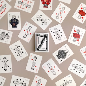 Könnte beinhalten: Ein Kartenspiel Grand Prix Legendary Drivers in einer weißen Schachtel mit schwarzem Text und Grafiken, umgeben von verstreuten einzelnen Spielkarten mit Illustrationen von Rennfahrern in roter, schwarzer und weißer Tinte auf weißem Karton.