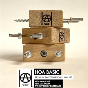Op de afbeelding: Een set van drie houten wandbeugels voor skateboards. De beugels zijn gemaakt van echt eikenhout en zijn ontworpen om skateboards op afstand van de muur te houden. De beugels zijn handgemaakt en passen op elke maat skateboard. De tekst op het label luidt "HOA BASIC Premium Skateboard Wandbeugel 100% handgemaakt Echt eikenhout Past op elke maat skateboard".