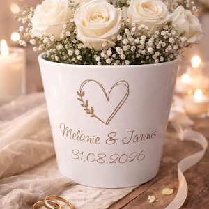Könnte beinhalten: Ein weißer Keramiktopf mit Herz- und Blättermotiv, gefüllt mit weißen Rosen und Schleierkraut. Auf dem Topf stehen die Namen "Melanie & Jannis" und das Datum "31.08.2026". Zwei goldene Eheringe befinden sich im Vordergrund.