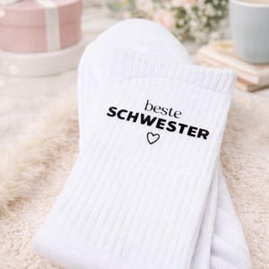Könnte beinhalten: Weiße Socken mit dem schwarzen Text "beste SCHWESTER" und einem Herzsymbol. Die Socken sind auf einer weichen, strukturierten Oberfläche gefaltet. Eine Geschenkbox und Blumen befinden sich im Hintergrund.