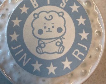 May include: A white ceramic mug with a blue and white sticker featuring the text "BTS" and a cartoon character. The sticker also includes stars and the text "Jin" and "RJ".