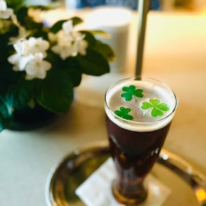 Op de afbeelding: Een glas donker bier met drie groene klavertjevier-stickers op de schuimkraag. Het glas staat op een gouden dienblad met een witte servet.