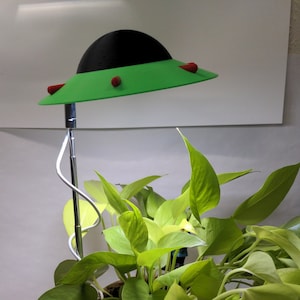 Op de afbeelding: Een groene en zwarte UFO-vormige lamp met rode accenten is geplaatst boven een potplant met lichtgroene bladeren.