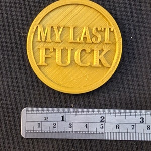 May include: A round, gold-colored object with the text "MY LAST FUCK" in bold, raised letters. The object is approximately 2 inches in diameter and has a textured surface. A ruler is placed below the object.