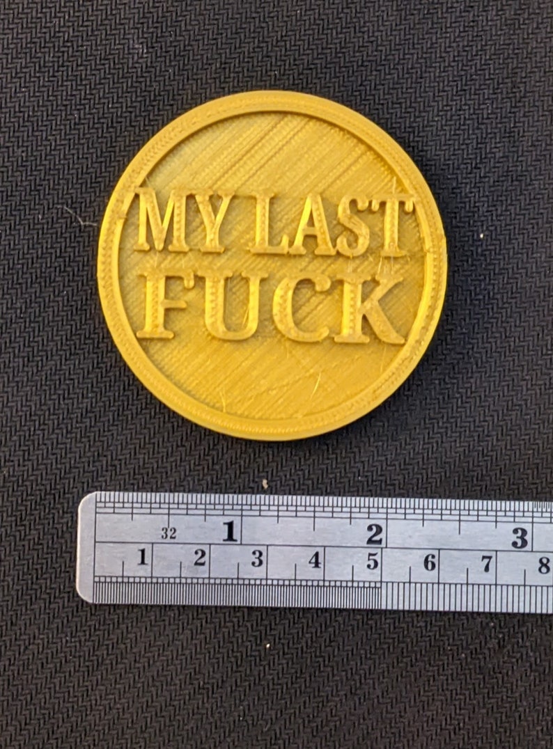 May include: A round, gold-colored object with the text "MY LAST FUCK" in bold, raised letters. The object is approximately 2 inches in diameter and has a textured surface. A ruler is placed below the object.