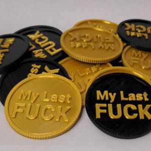 May include: A collection of black and gold coins. Each coin has the phrase "My Last FUCK" embossed on it. The coins are scattered across a light-colored surface, with some coins overlapping.