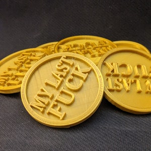 May include: A collection of gold-colored, circular tokens. Each token has the phrase "MY LAST FUCK" embossed in bold, capital letters. The tokens are arranged in a slightly overlapping pattern, with a textured background.