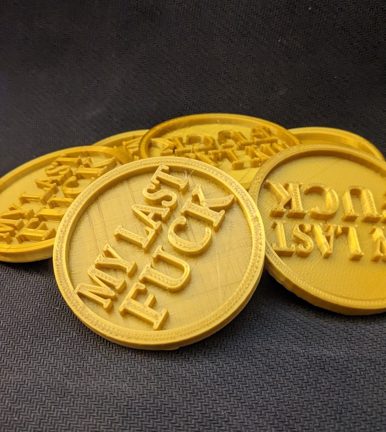 May include: A collection of gold-colored, circular tokens. Each token has the phrase "MY LAST FUCK" embossed in bold, capital letters. The tokens are arranged in a slightly overlapping pattern, with a textured background.