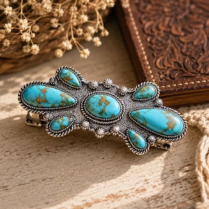 May include: A silver-toned hair clip with turquoise-colored stones and a decorative design. The clip features multiple oval and teardrop-shaped stones with brown veining. The metal has a detailed, textured border.