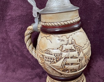 May include: A ceramic beer stein with a pewter lid and a rope-like handle. The stein features a detailed image of a ship sailing into a harbor with buildings in the background. The text "Keith Barkentine" is visible on the base.