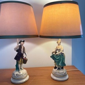 May include: Two table lamps with porcelain figurines. The lamp on the left features a man in a brown and green outfit holding a basket. The lamp on the right features a woman in a green dress. Both lamps have beige lampshades with green trim.