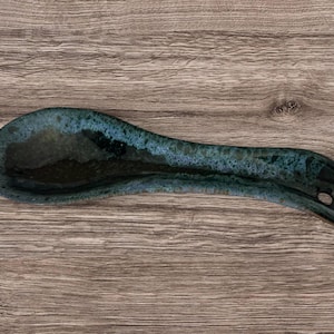 Könnte beinhalten: Eine Keramik-Löffelablage mit dunkler tealfarbener Glasur und einem kleinen Loch im Griff. Die Löffelablage ist oval geformt und liegt auf einer Holzoberfläche. Die Glasur hat ein gesprenkeltes, strukturiertes Aussehen.