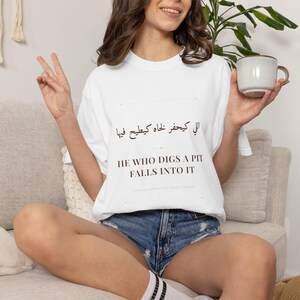 Peut inclure: T-shirt blanc avec l'inscription "HE WHO DIGS A PIT FALLS INTO IT" en anglais et en arabe. Le t-shirt est porté avec un short en jean. Une personne tient une tasse blanche.